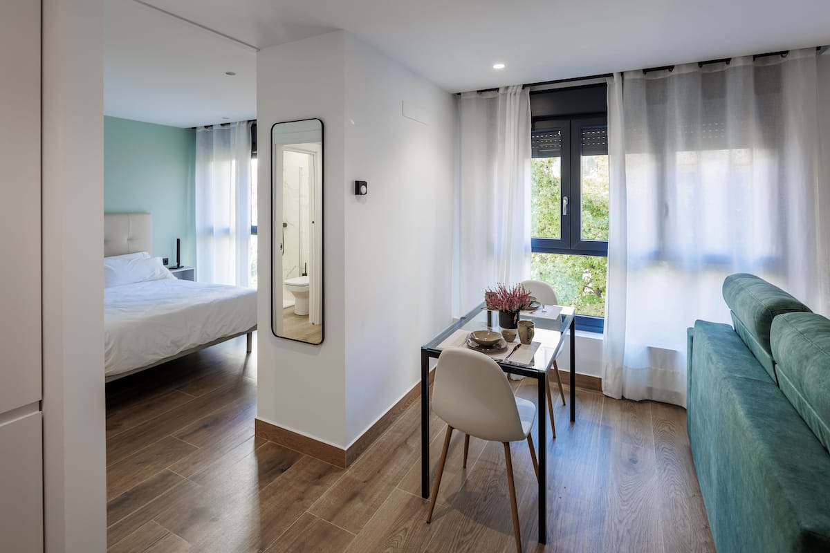 A modern room with a bed, a dining table with chairs, a sofa, and a large window with curtains.