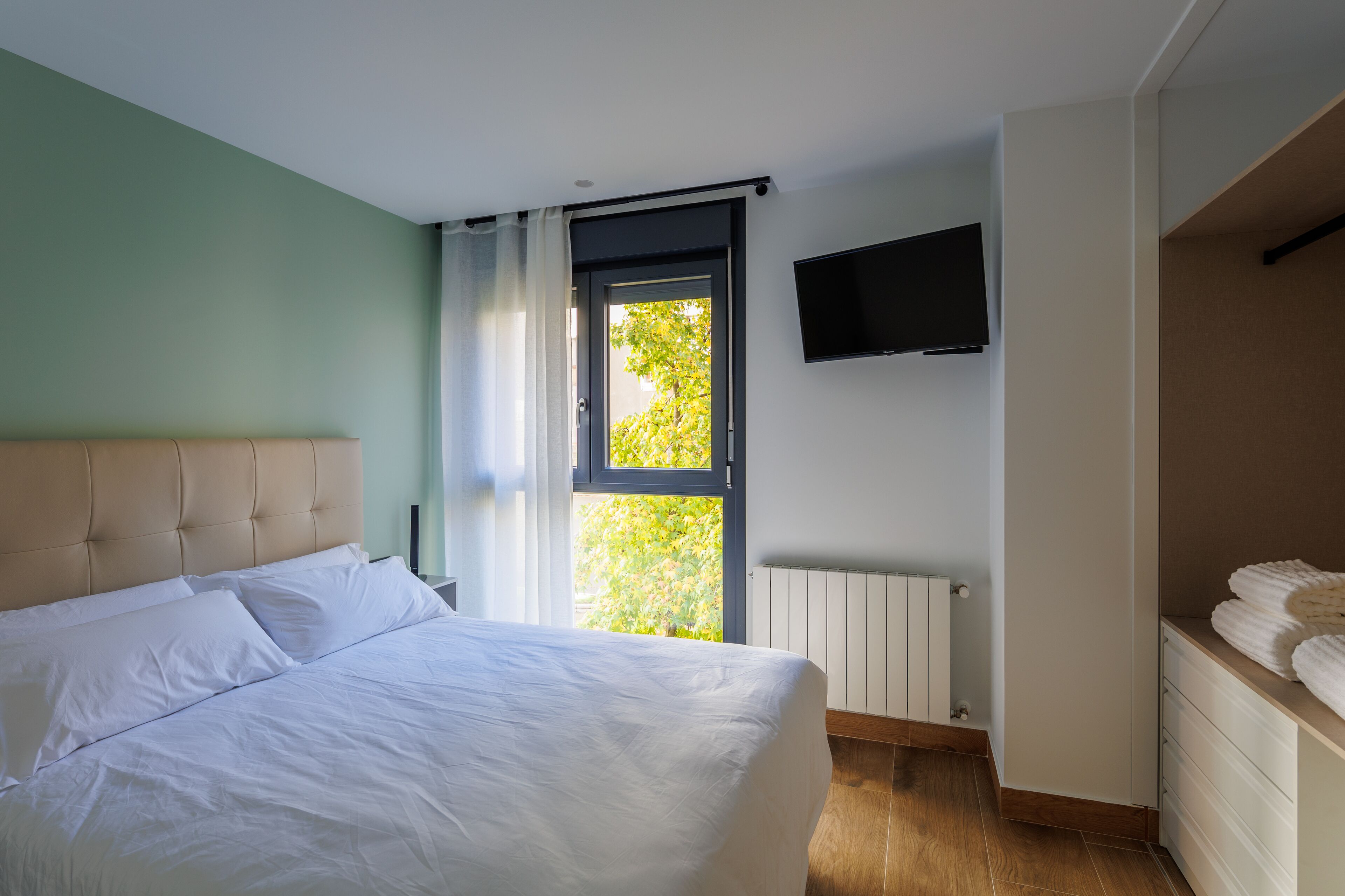 A bedroom with a bed, a window with curtains, a wall-mounted TV, and a radiator.