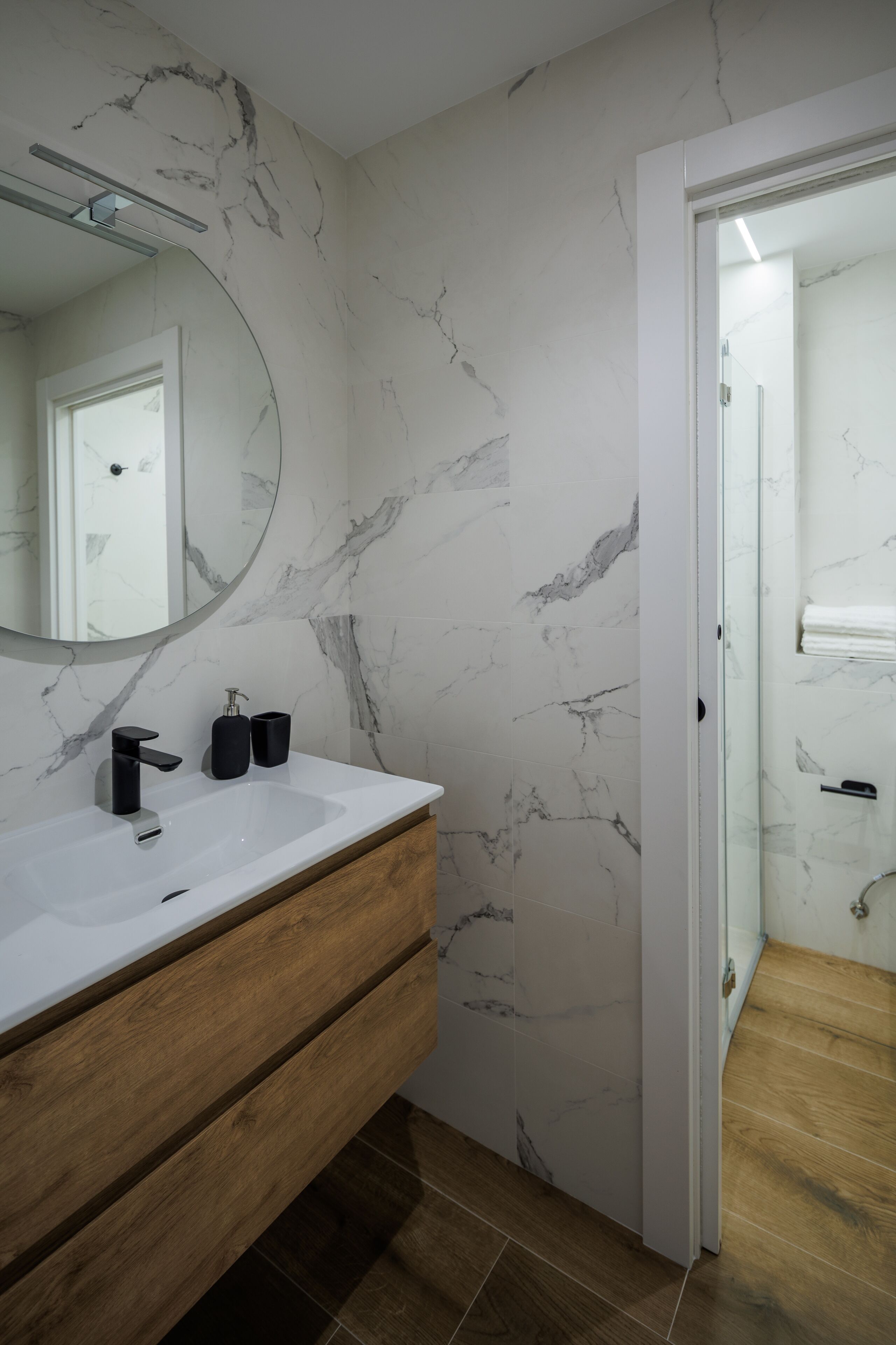 A modern bathroom with a marble backsplash, a wooden vanity, a large mirror, and a glass shower enclosure.