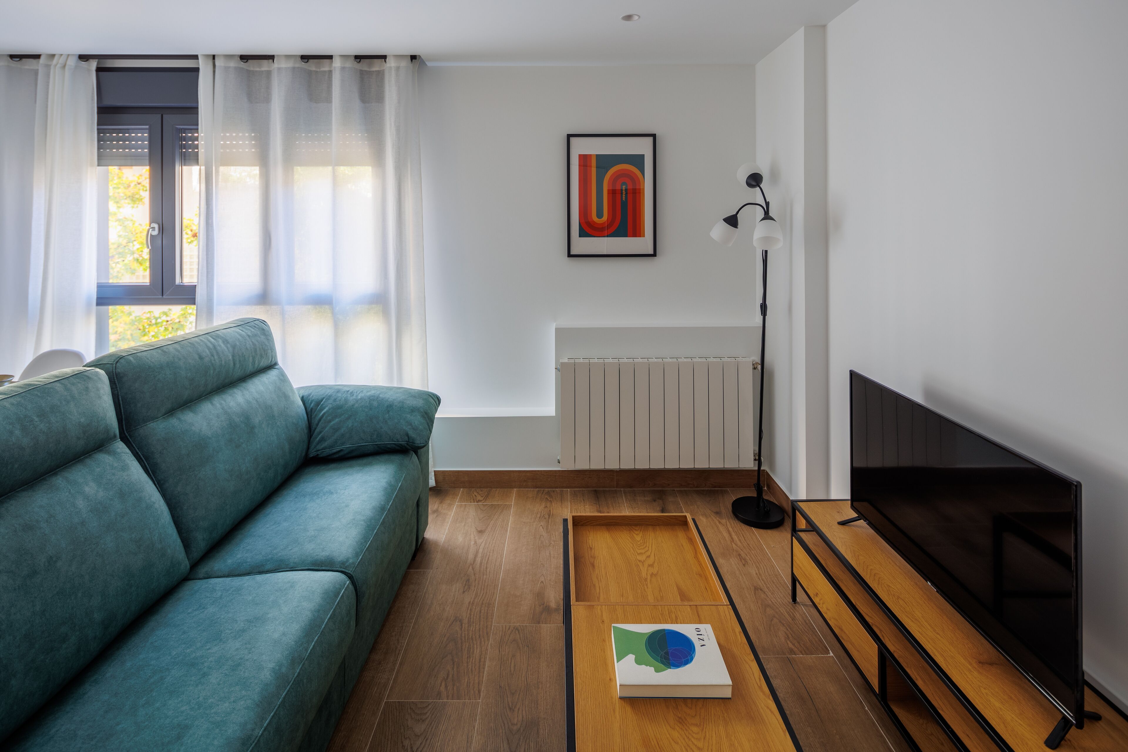Una sala de estar moderna con un sofá color turquesa, una mesa de centro de madera con un libro, un televisor de pantalla plana y un radiador.