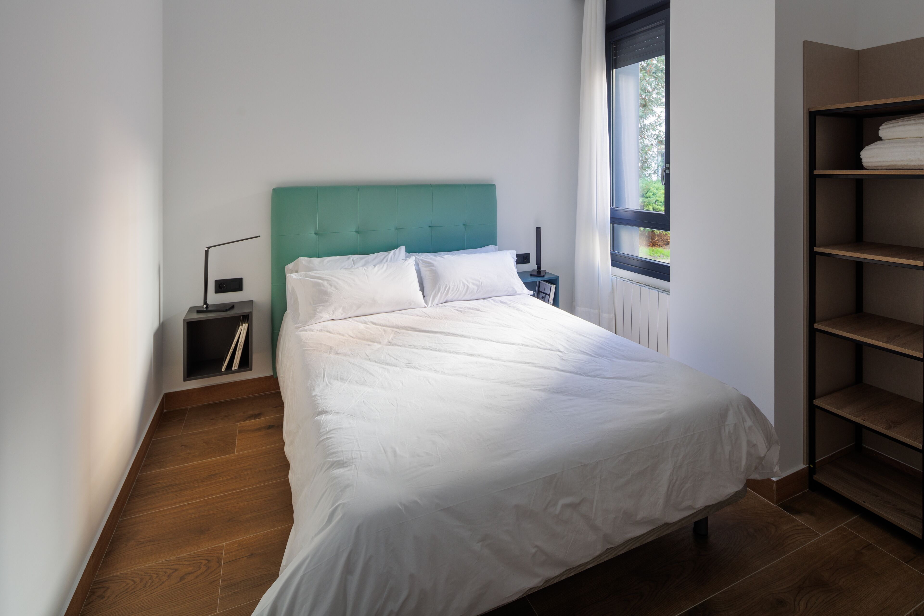 A bedroom with a bed, a nightstand, a lamp, a window, and a built-in shelving unit.