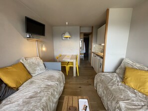 Habitación compacta con dos camas, televisor montado en la pared, mesa de comedor con sillas y kitchenette.