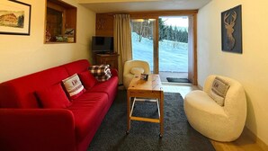 Ein gemütliches Wohnzimmer mit einem roten Sofa, einem weißen Sessel, einem Holztisch und einem Fernseher auf einem Schrank.