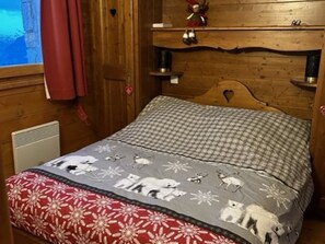 Ein hölzernes Bett mit einem gemusterten Bettbezug und Kissen, ein Fenster mit Ausblick und ein roter Vorhang.