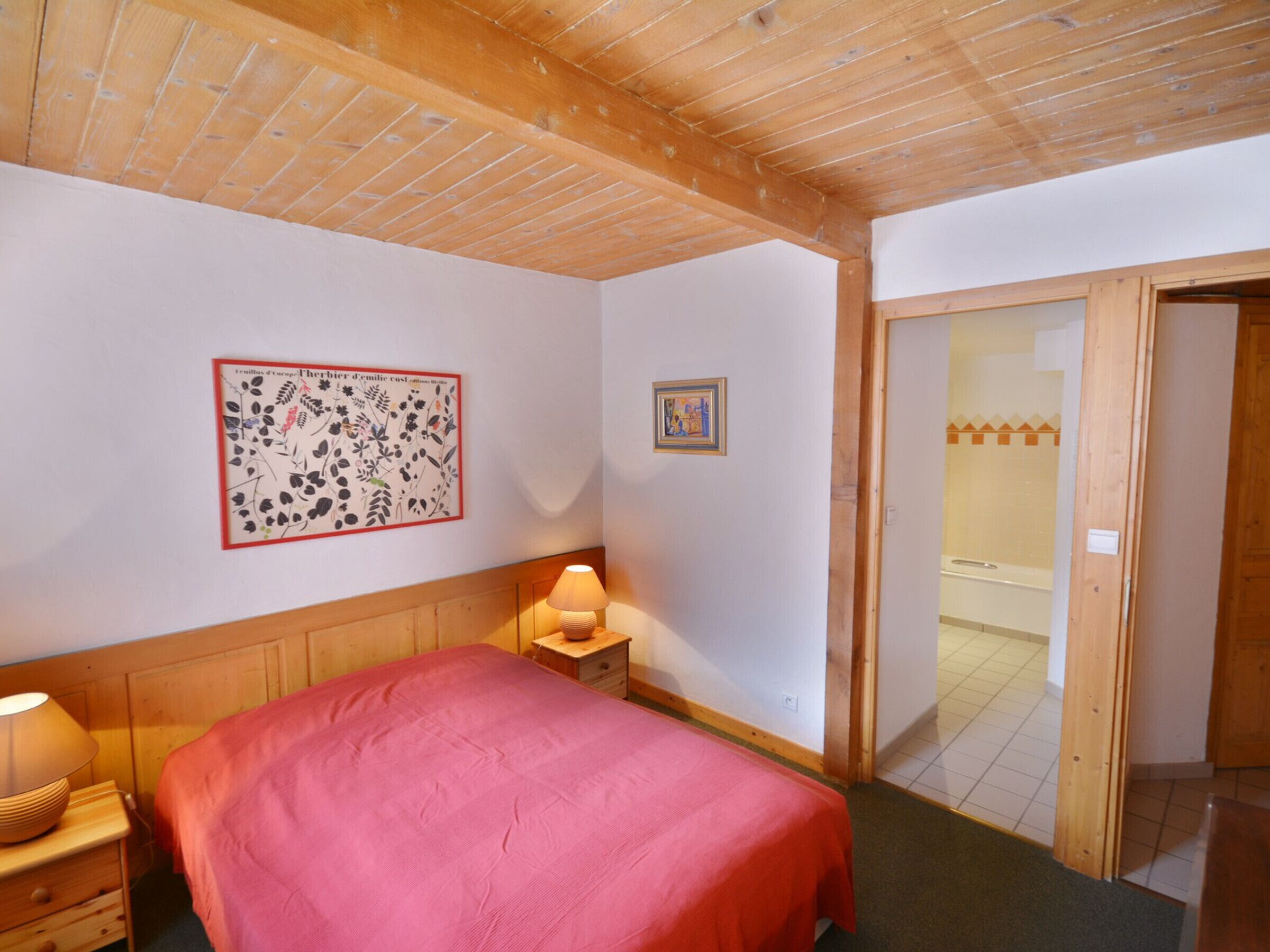 Une chambre avec un plafond en bois, un lit recouvert d’une couverture rouge, une table de chevet avec une lampe et une porte donnant sur une salle de bain.