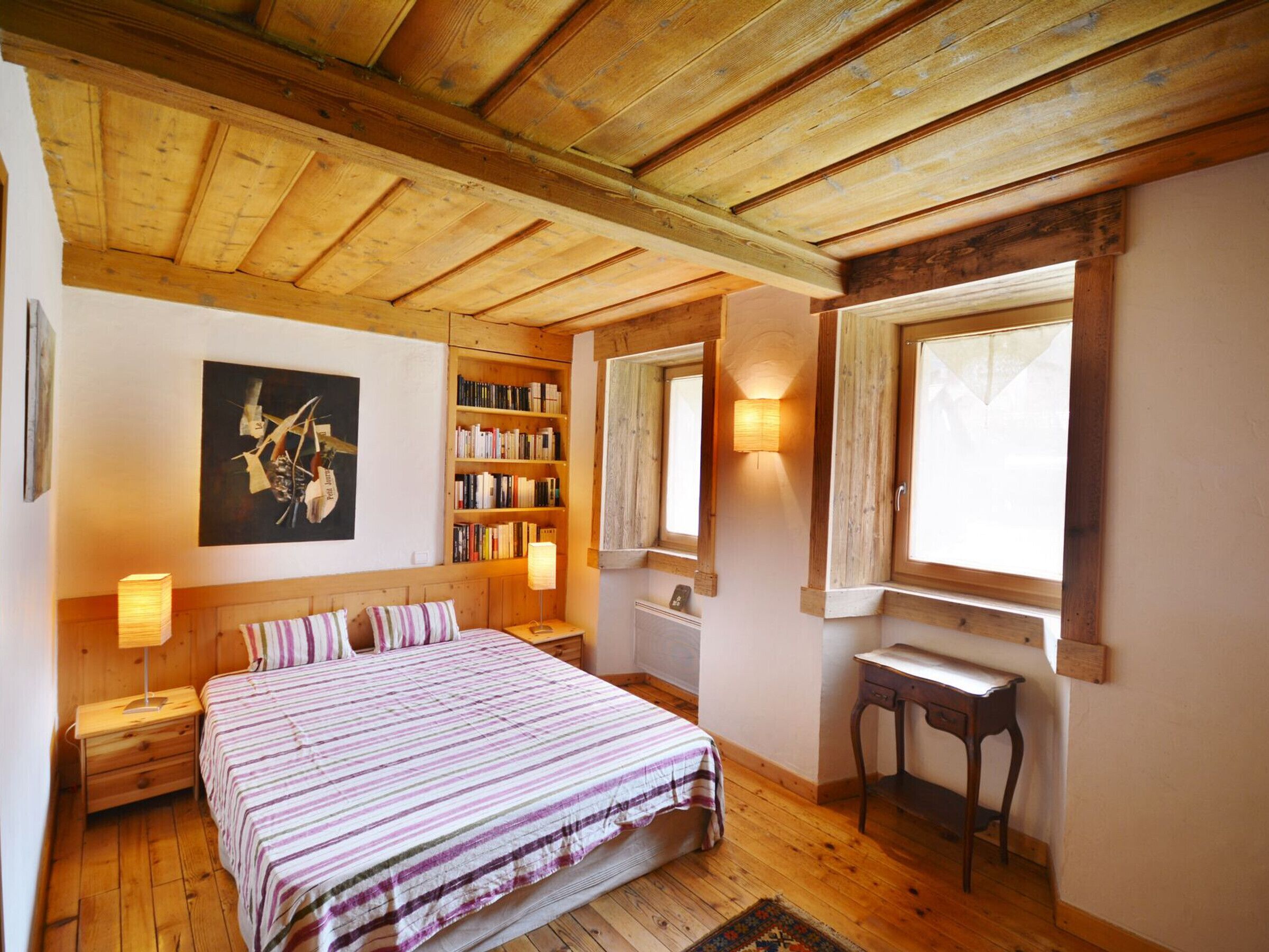 A cozy bedroom with a wooden ceiling, a large bed with striped bedding, a bedside table with a lamp, a bookshelf, and a window with a view.