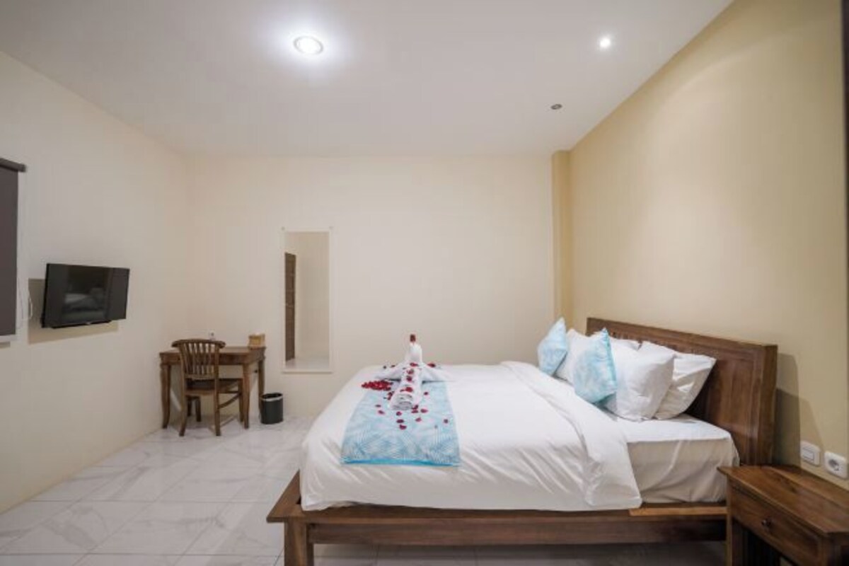 A bedroom with a bed, a wooden nightstand, a chair, a small table, and a TV mounted on the wall.