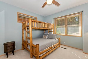 Una cama litera con estructura de madera, una mesita de noche pequeña y una ventana con persianas.