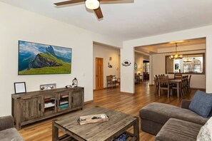 A spacious living room with a large flat-screen TV, wooden entertainment unit, and a dining area with a chandelier.