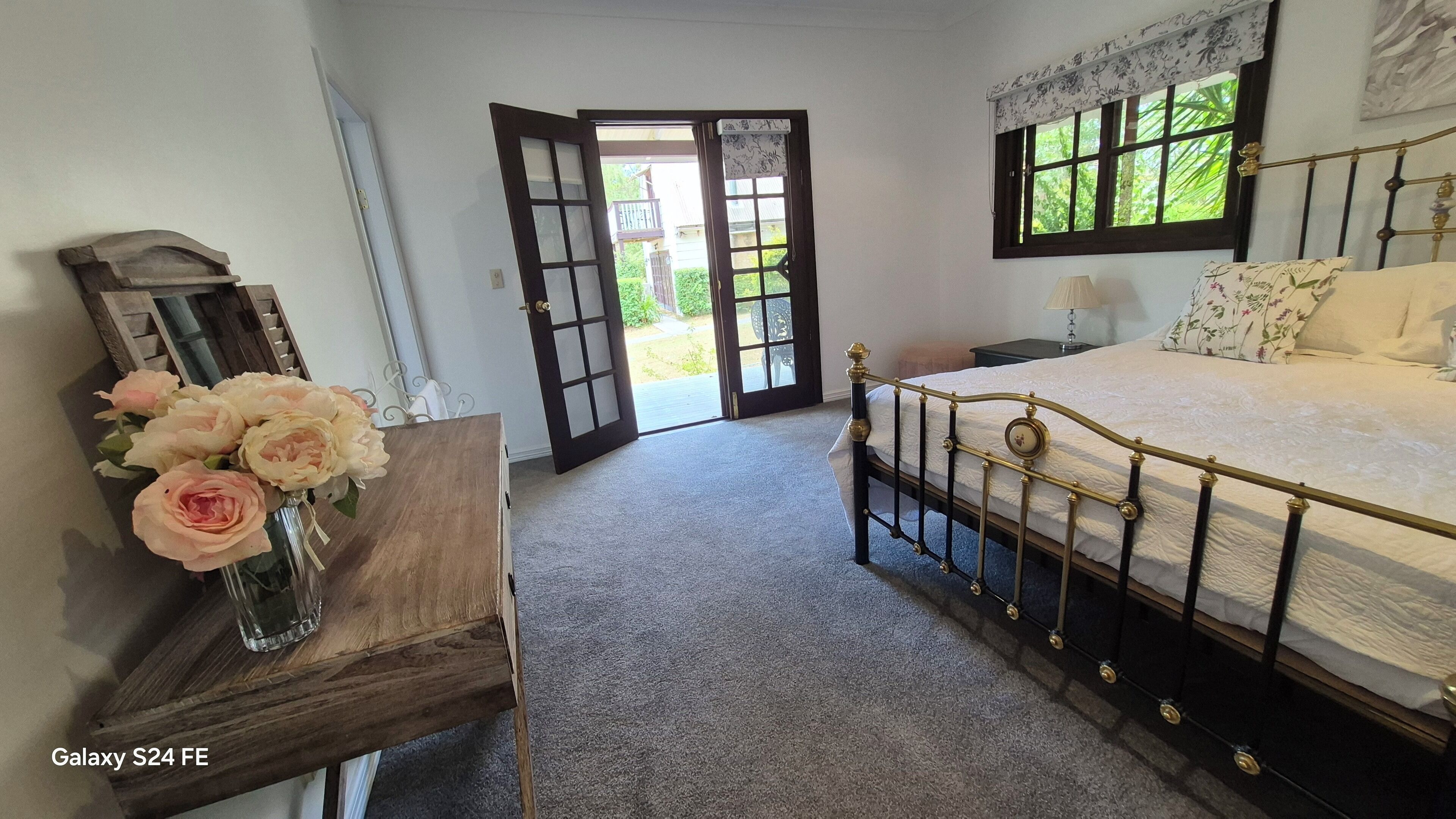 Een slaapkamer met een bed, een houten nachtkastje, een vaas met bloemen en een deur naar buiten.