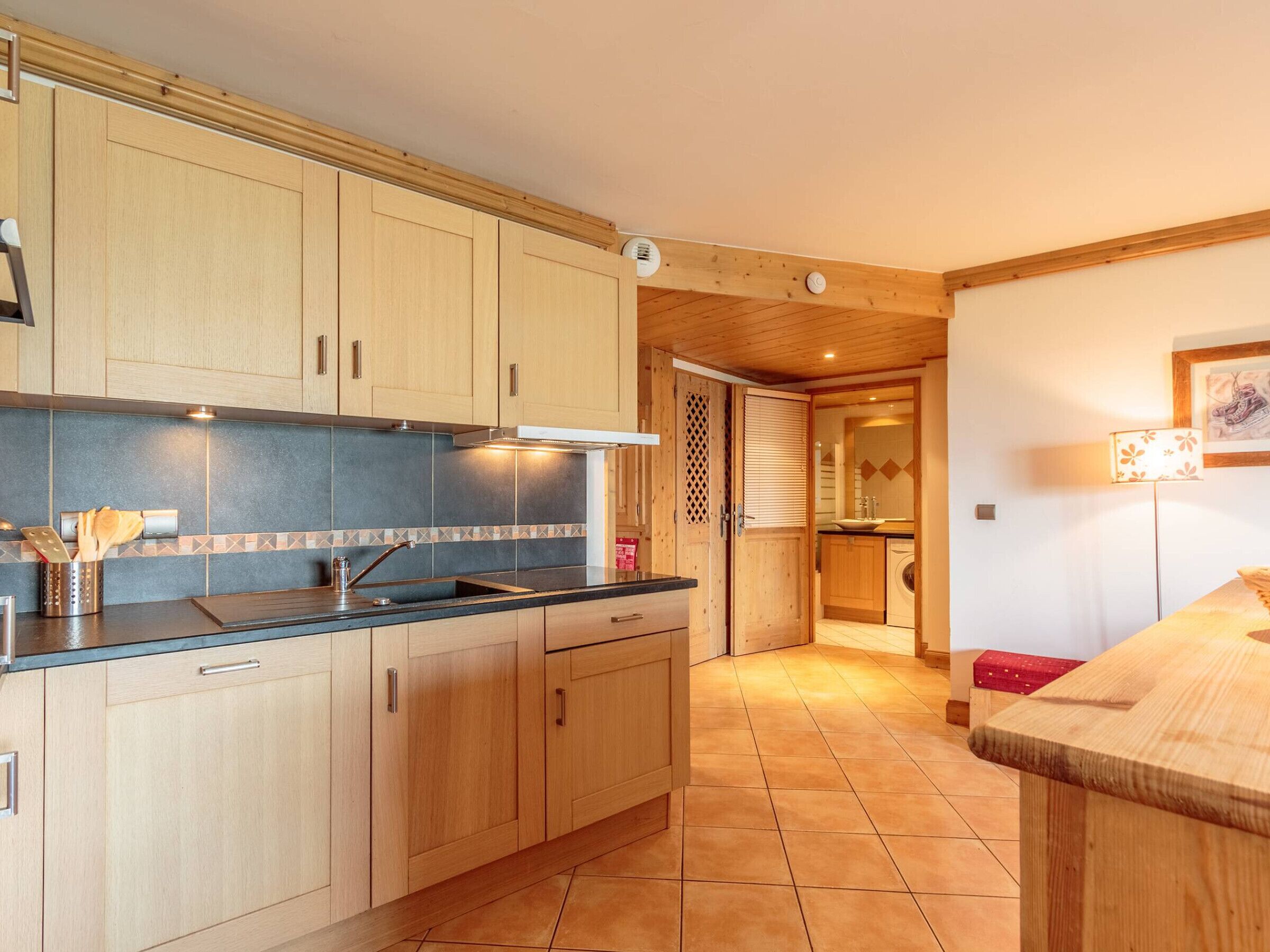 A kitchen with wooden cabinets, a sink, and a countertop. There is a window with blinds and a door leading to a bathroom.