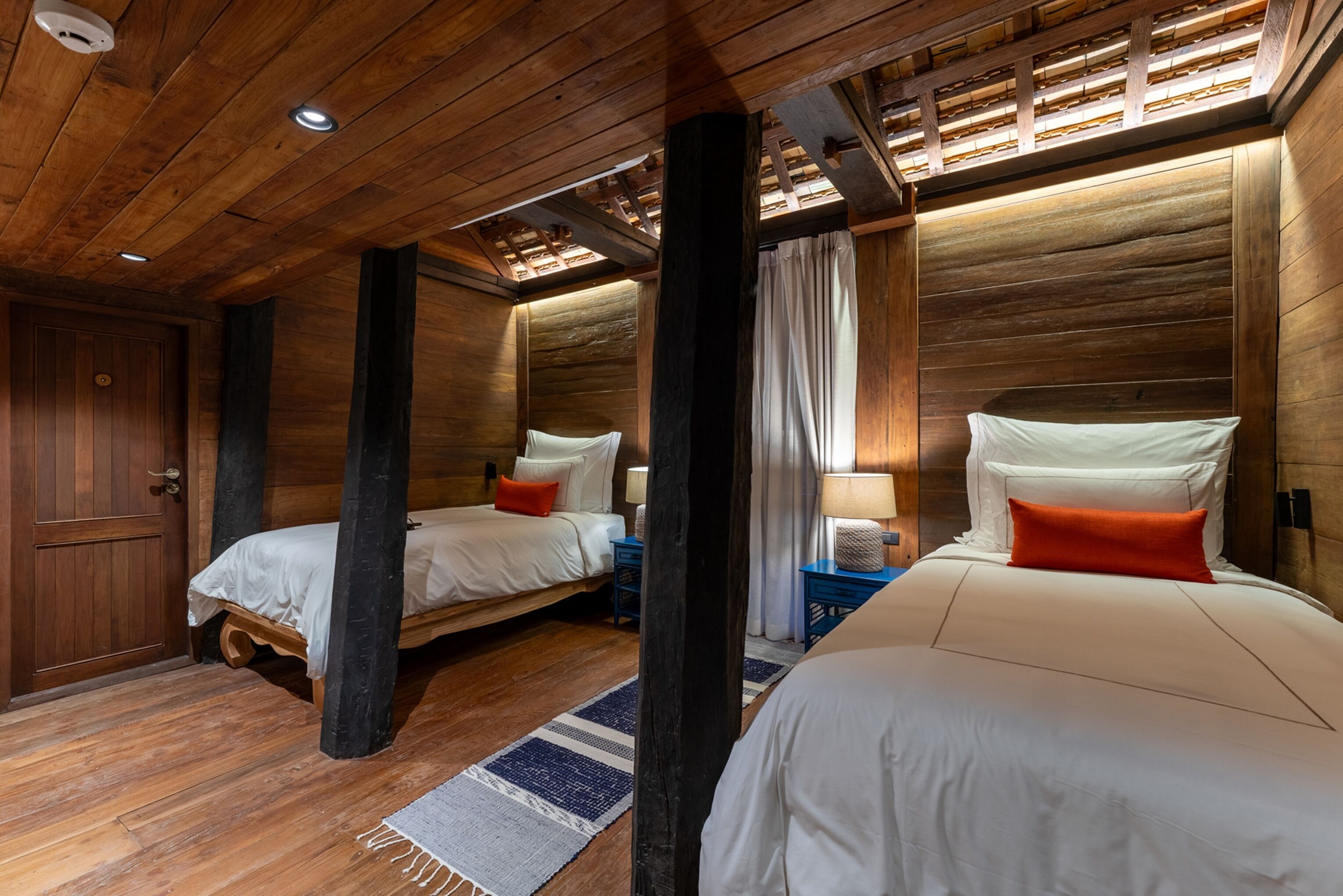 A wooden cabin-style bedroom with two beds, a wooden door, and a window with curtains.