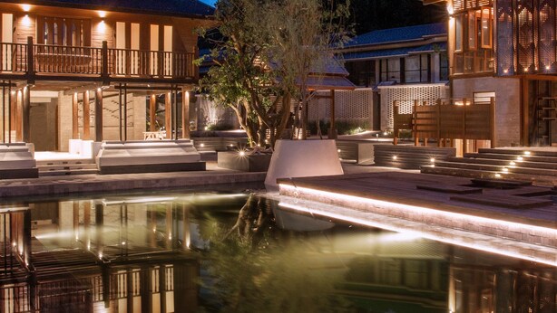 Ruhiger Poolbereich mit Spiegelung der Gebäude, einer Holzterrasse und einem Nachthimmel mit Mond.