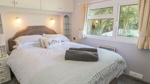 A neatly made bed with pillows and a blanket, a bedside table with a lamp, and a window with curtains. - Orchard Lodge (Pwllheli)