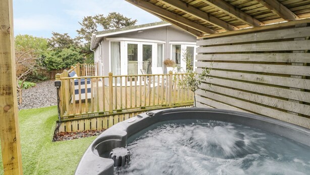 A hot tub under a wooden pergola, a fenced patio area, and a white building with glass doors. - Orchard Lodge (Pwllheli)