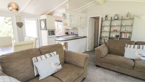 A spacious living area with two sofas, a kitchen with white cabinets, and a bookshelf filled with books and decorative items. - Orchard Lodge (Pwllheli)