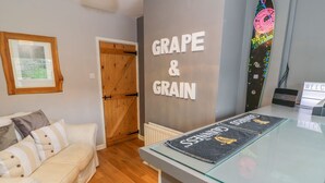 Una stanza con un divano bianco, una porta in legno, un muro con la scritta 'GRAPE & GRAIN' e un tavolino in vetro con un sottobicchiere.