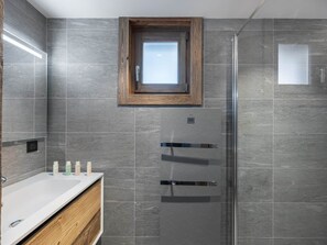 A modern bathroom with grey tiles, a wooden medicine cabinet, a glass shower enclosure, and a small window above the sink.
