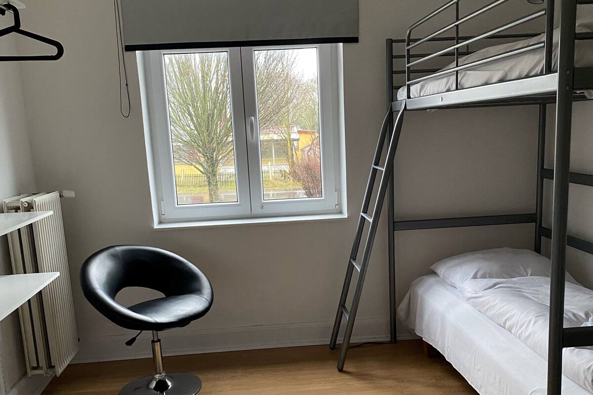 Une chambre de dortoir avec des lits superposés, un radiateur, une chaise et une fenêtre donnant sur des arbres et une clôture.