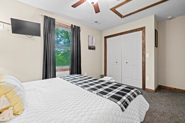 A bedroom with a bed, a ceiling fan, a window with curtains, a door, and a flat-screen TV mounted on the wall. - Silver Linings - Hot Tub, Mountain Views, Close to Town, Pet Friendly (Waynesville)