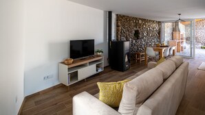 Ett modernt vardagsrum med en platt-TV, ett svart kylskåp, en vägg av sten, en matplats med bord och stolar samt ett stort fönster.