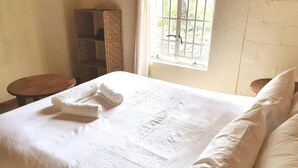 Un lit bien fait, avec du linge de lit blanc et une serviette enroulée, une chaise en bois et une fenêtre avec des barreaux.