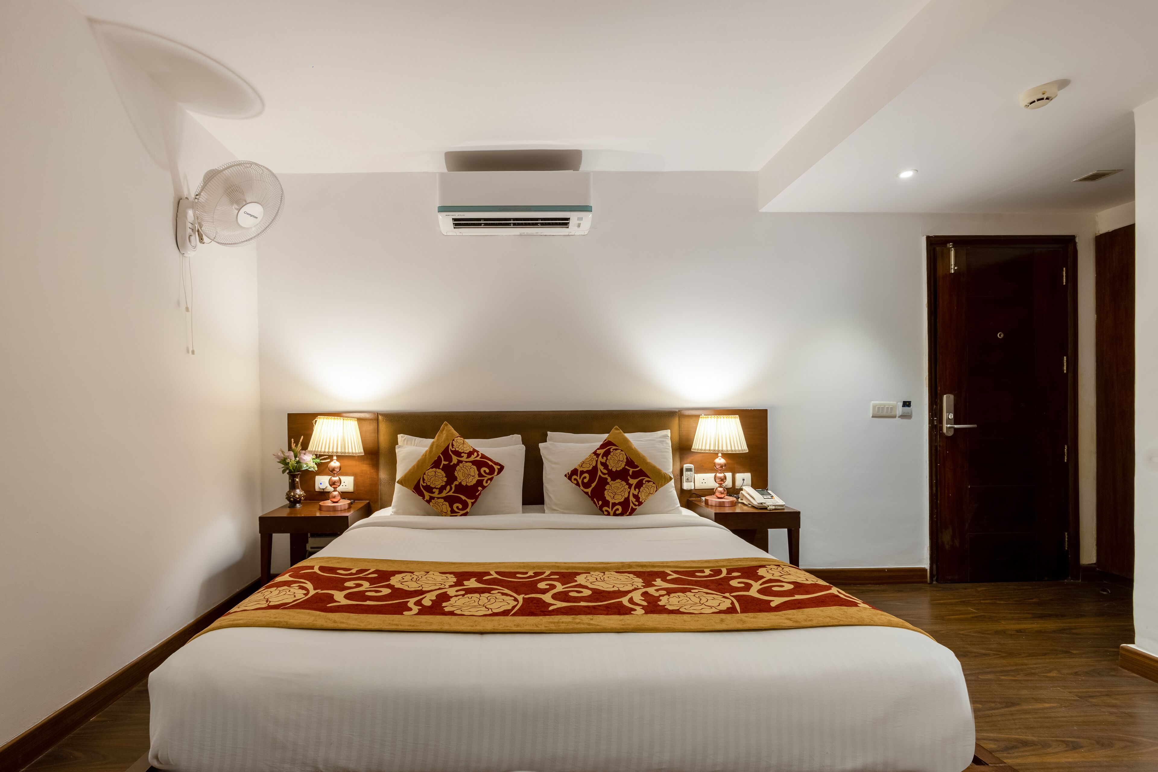 A hotel room with a large bed, two bedside tables with lamps, an air conditioning unit, and a wooden door.