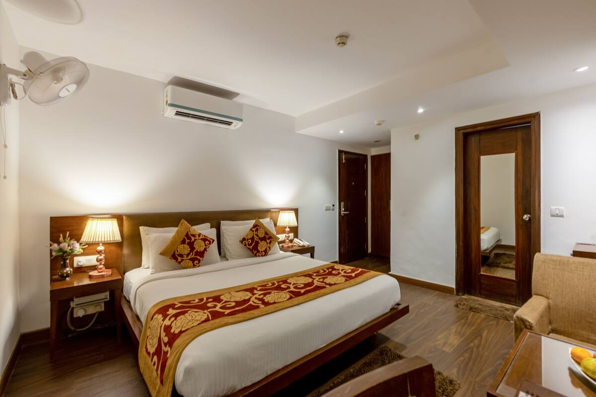 Une chambre d’hôtel avec un grand lit, des lampes de chevet, une tête de lit en bois, un appareil de climatisation, une porte en bois et une petite table sur laquelle se trouve un panier de fruits.