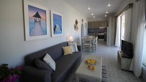 Un salon moderne avec un canapé, une table basse et des œuvres d’art accrochées au mur. On aperçoit à l’arrière-plan un coin cuisine avec une table à manger et des chaises.