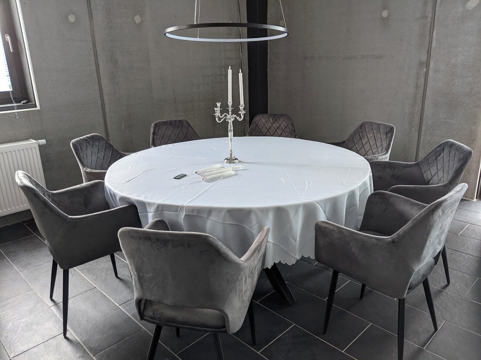 A round table with a white tablecloth, surrounded by six grey upholstered chairs with black legs. A candelabra with candles is placed on the table.