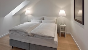 Een moderne slaapkamer met een schuine wand, een bed met witte lakens, twee bedlampjes en een lijst met een afbeelding aan de muur.