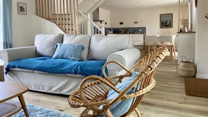 A modern living room with a sofa, wicker chairs, a staircase, and a kitchen in the background. - Charming house in the port of Sainte-Marine (Combrit)