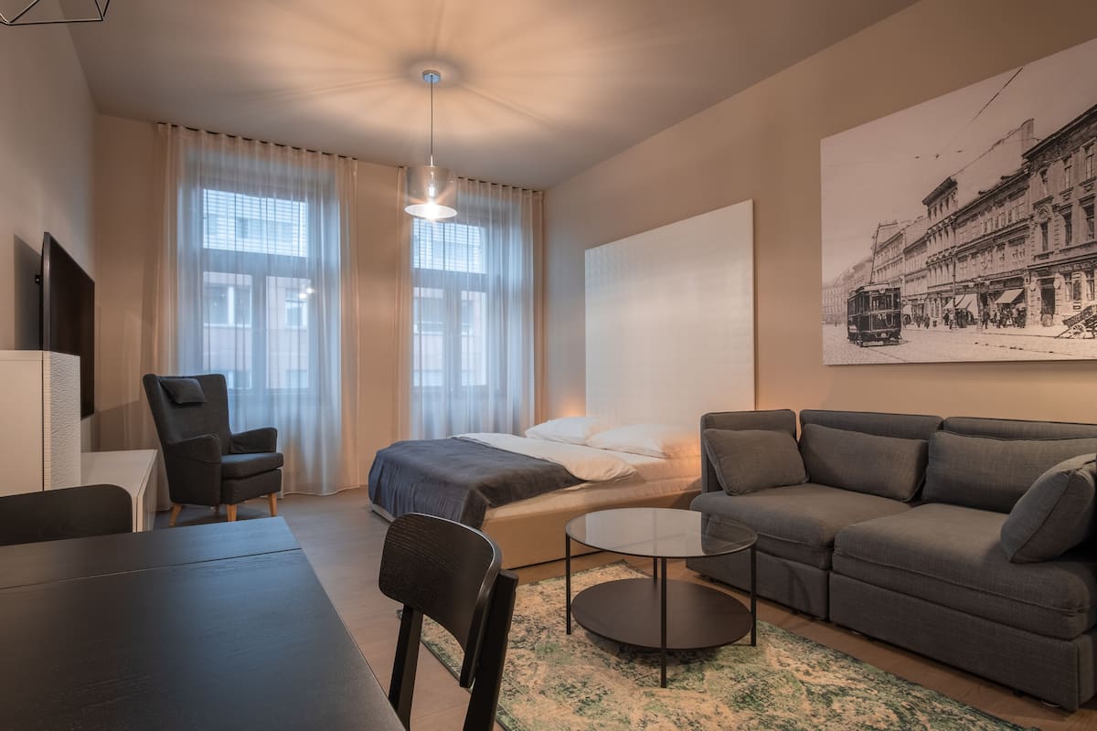 A modern hotel room with a bed, a sofa, a dining table, and a TV.