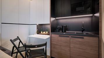 Una cocina moderna con encimera blanca, pileta negra y muebles de madera. Se observa una silla y una pequeña mesa con botellas encima.