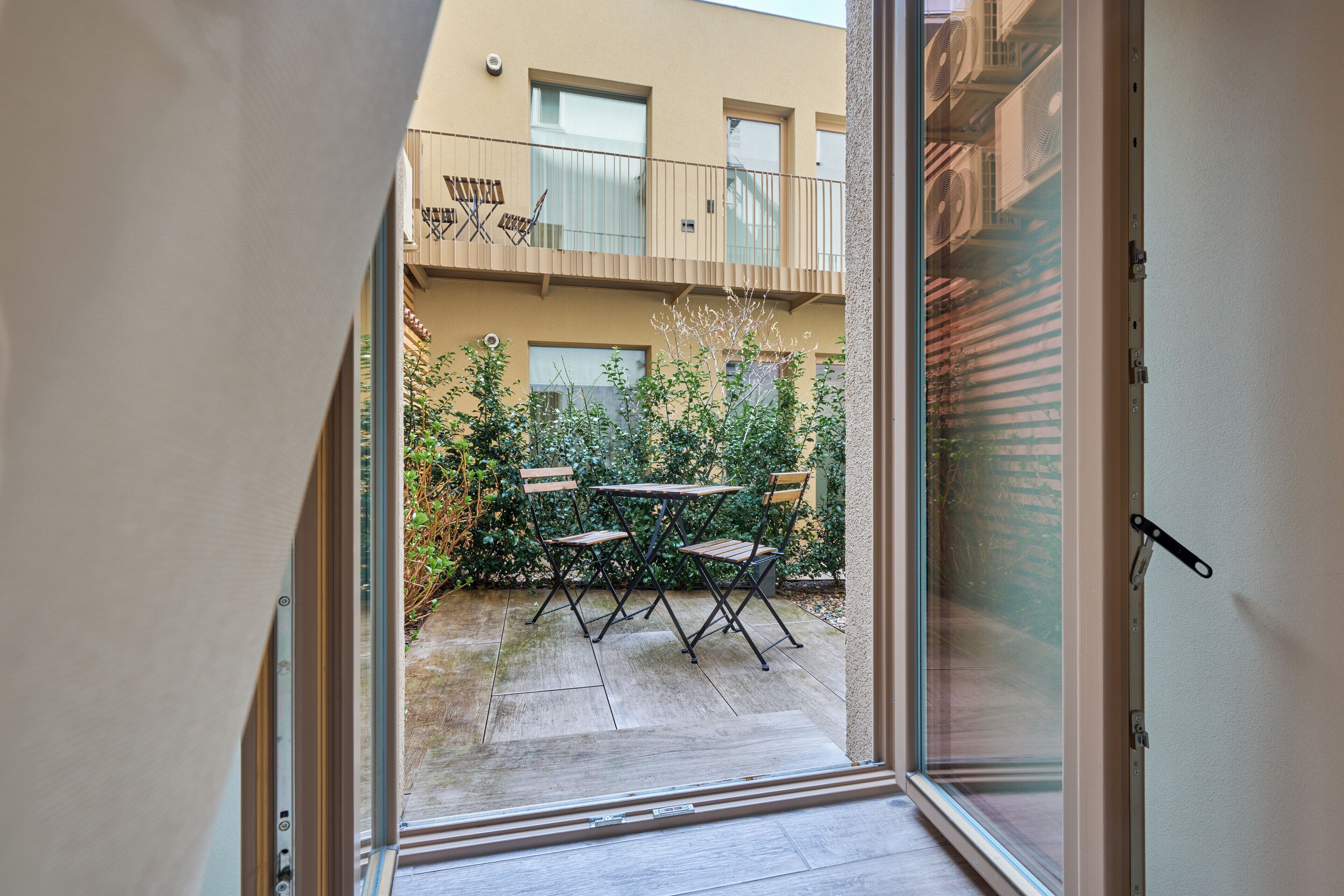 Puerta corrediza abierta que da a un patio al aire libre con una mesa y sillas, rodeado de follaje.