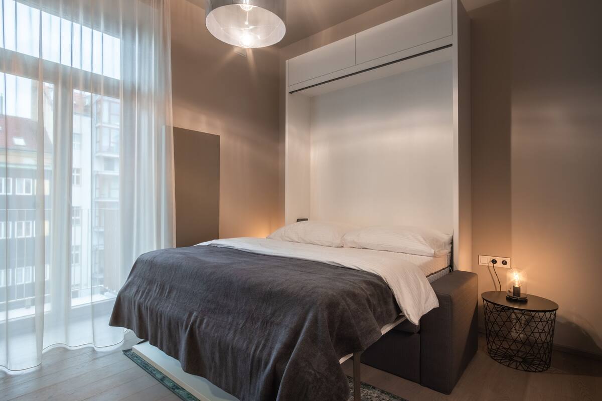 A modern bedroom with a bed, a bedside table with a lamp, and a view of the city through sheer curtains.