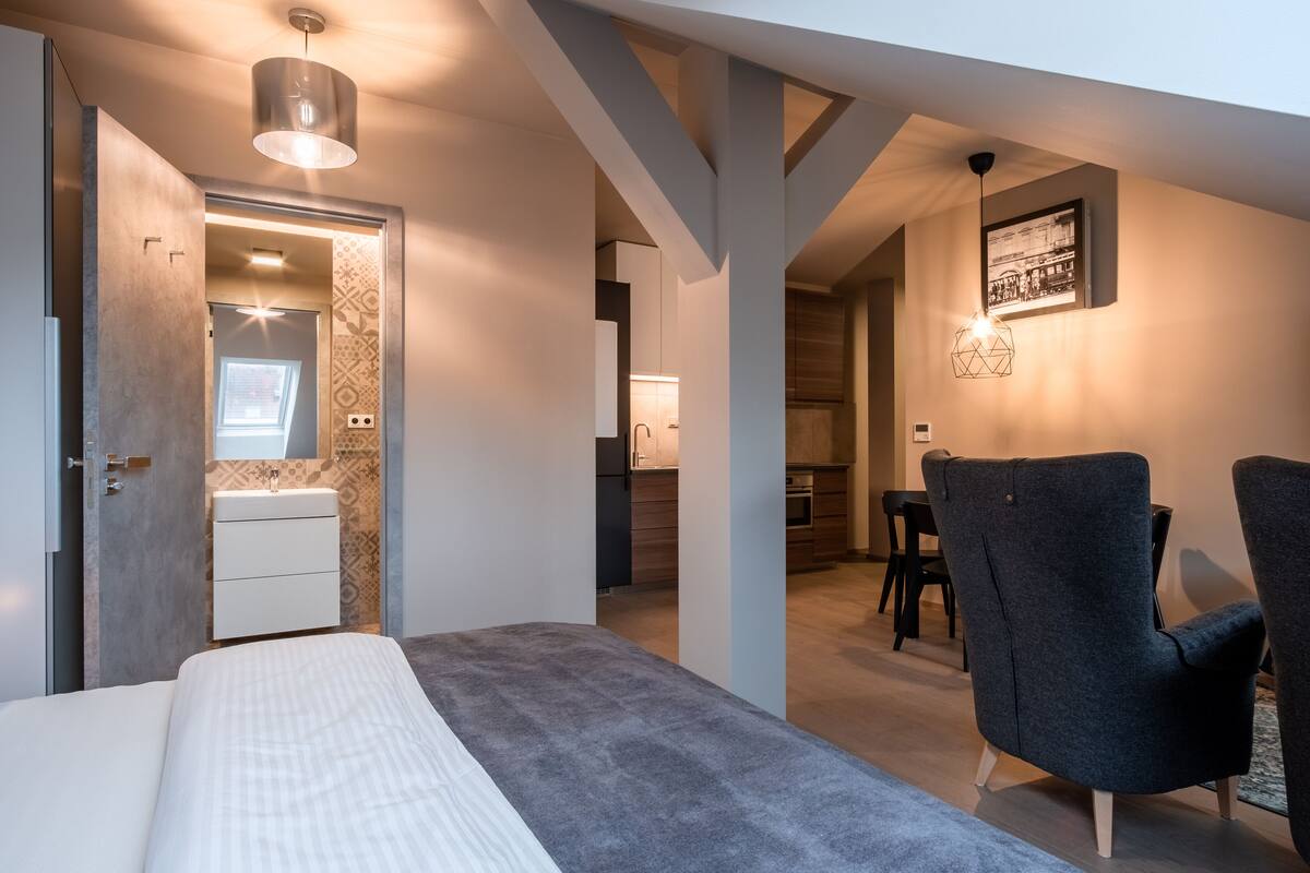 A modern bedroom with a bed, a nightstand, a dresser, and a bathroom with a sink and mirror. There is a dining area with a table and chairs, and a kitchen in the background.