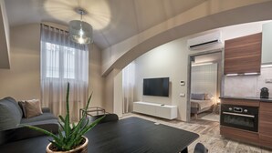 Una sala moderna con una mesa de comedor de madera oscura, un sofá y una planta grande en maceta. Hay un televisor montado en la pared y una zona de cocina con horno y microondas incorporados.