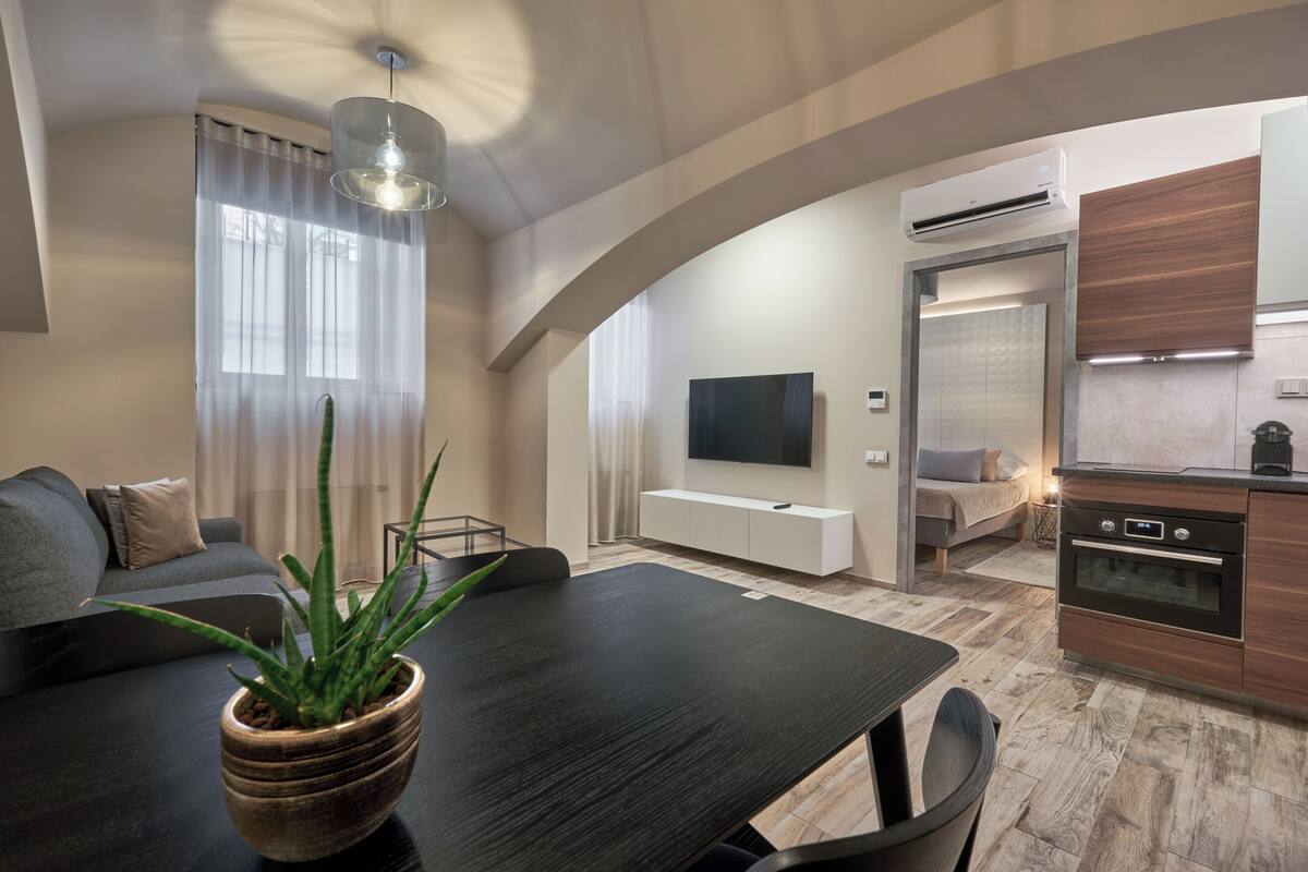 A modern living room with a dark wooden dining table, a sofa, and a large potted plant. There is a TV mounted on the wall and a kitchen area with a built-in oven and microwave.