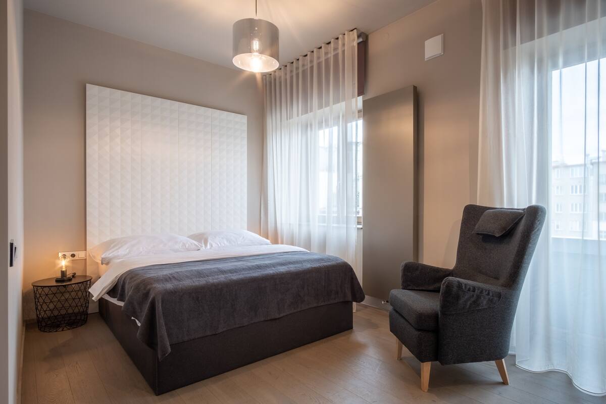 A modern hotel room with a large bed, a side table with a lamp, a chair, and a large window with sheer curtains.