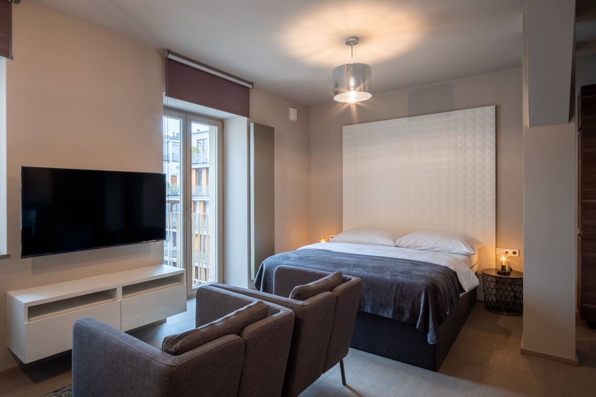 A modern bedroom with a flat-screen TV, a bed with a white headboard, a bedside table with a lamp, and a window with a view of buildings.