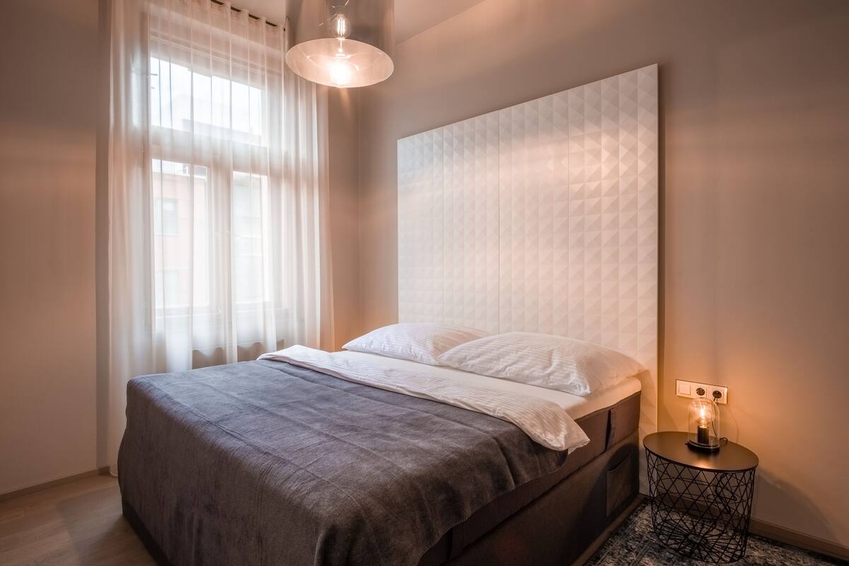 A neatly made bed with a grey bedspread and white pillows, a round bedside table with a lamp, and sheer curtains on a window.