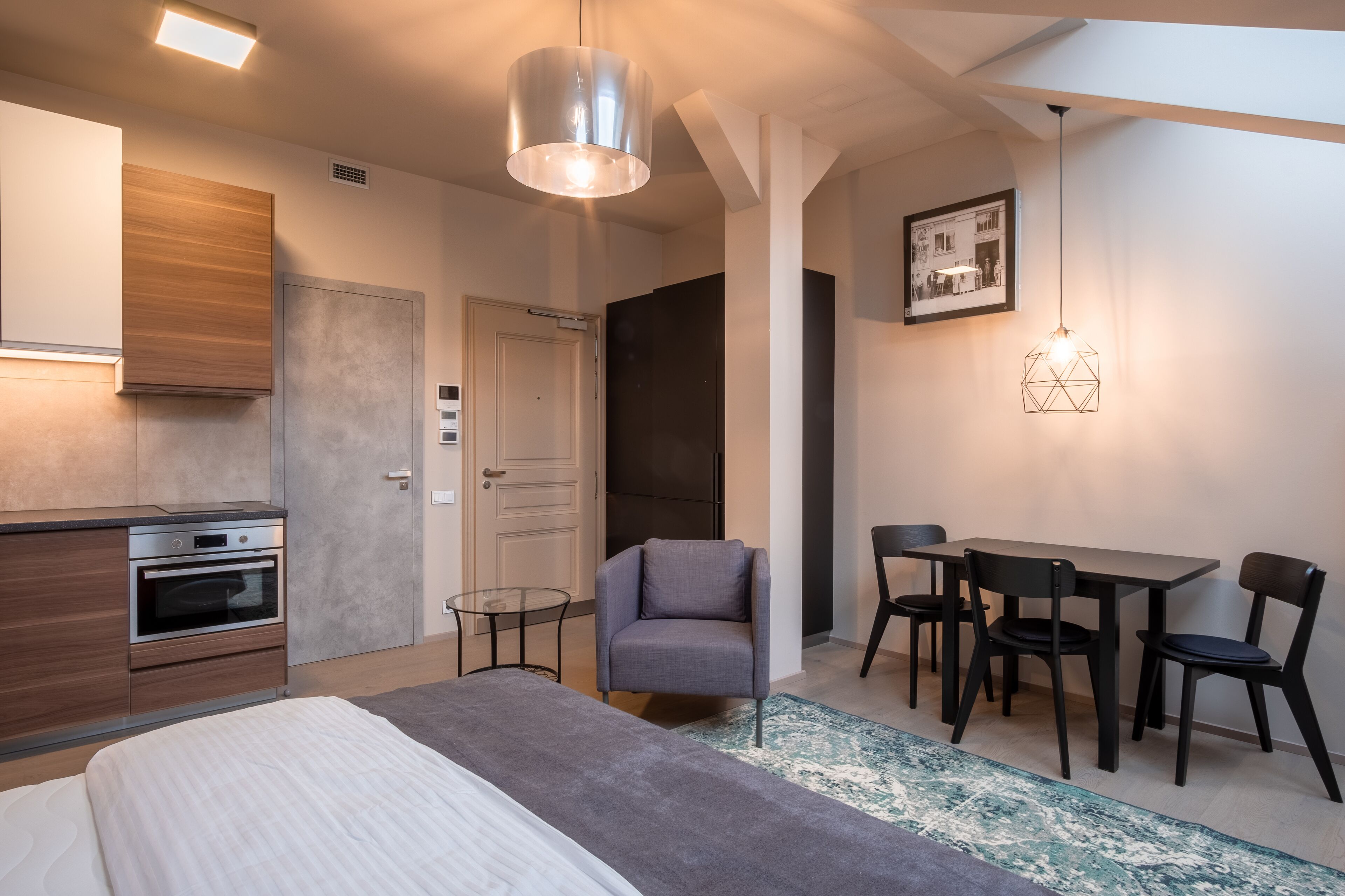 A modern hotel room with a bed, a desk with chairs, a kitchenette, and a dining area.