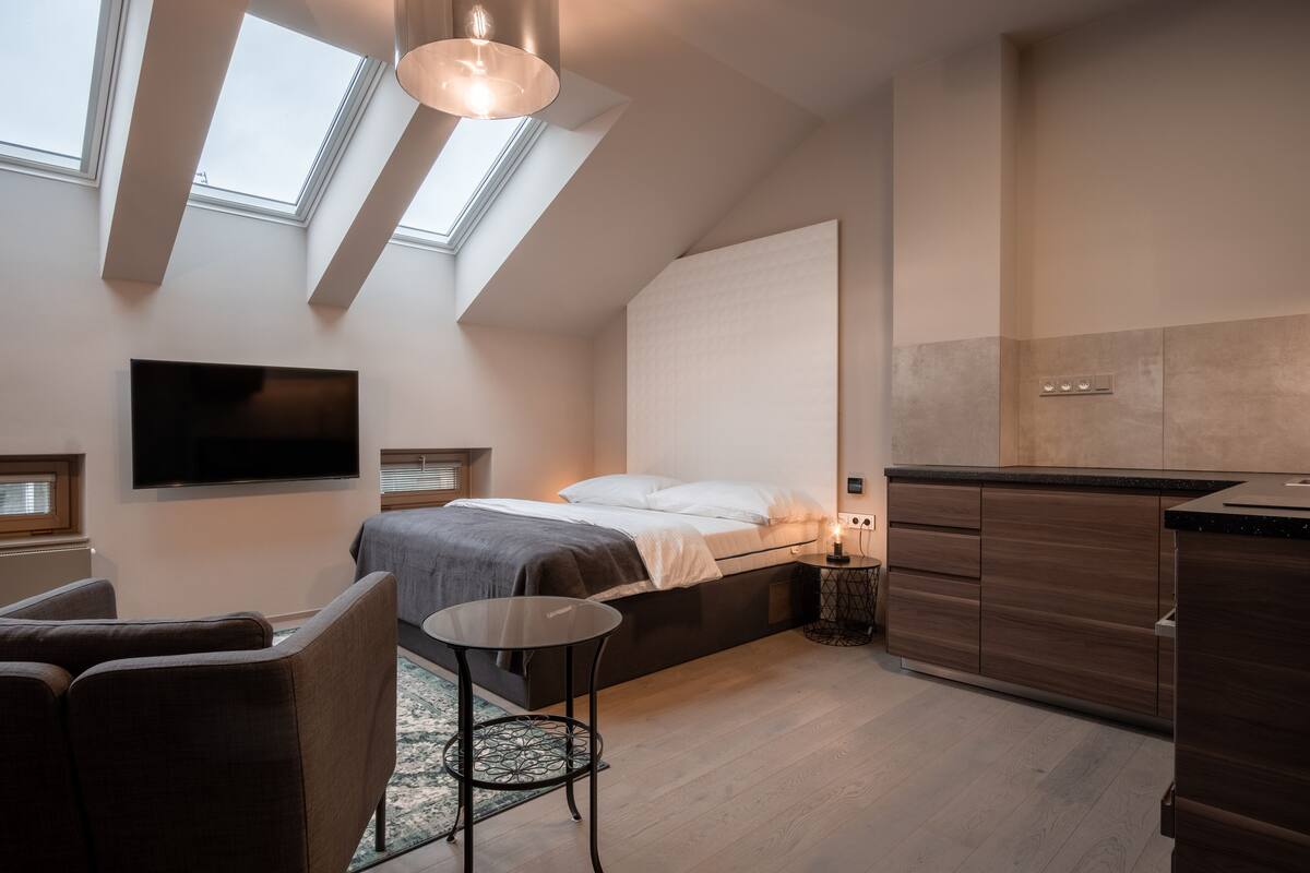 A modern bedroom with a bed, a TV, a small table, and a sofa. There are skylights and a kitchen area visible.
