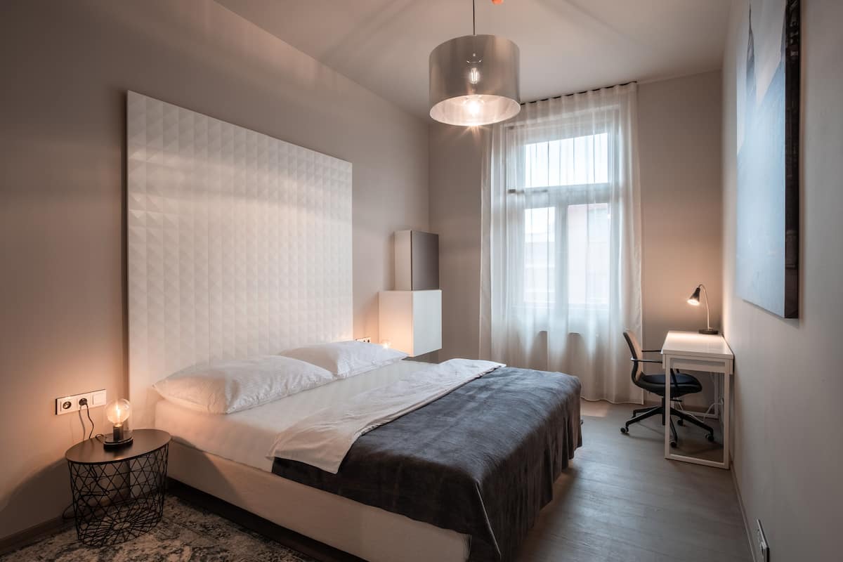 A modern bedroom with a large bed, bedside table with a lamp, a desk with a chair, and a large window with sheer curtains.