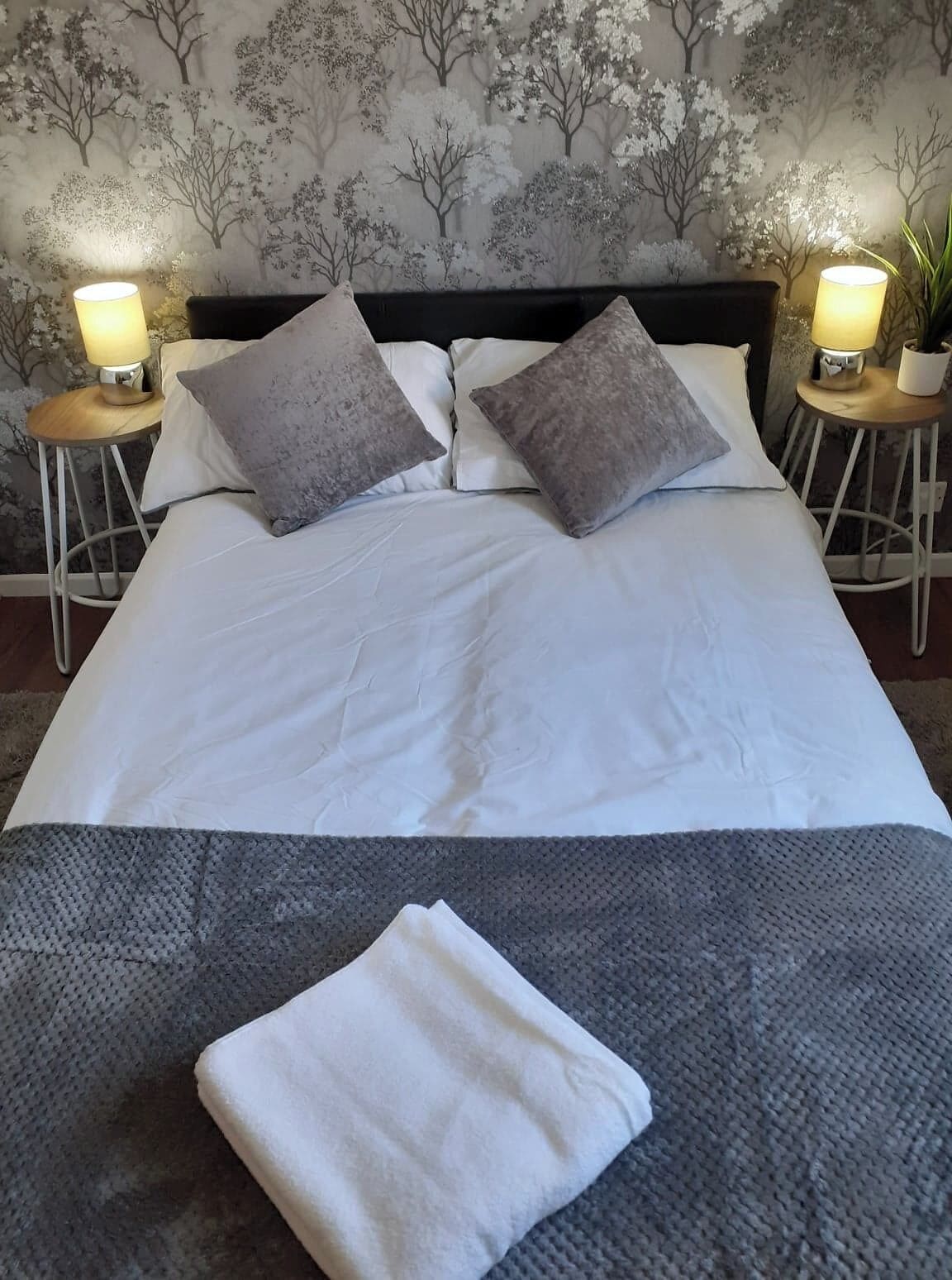 Una cama bien hecha con sábanas blancas y dos almohadas grises, con dos lámparas de noche a cada lado sobre mesitas a juego.