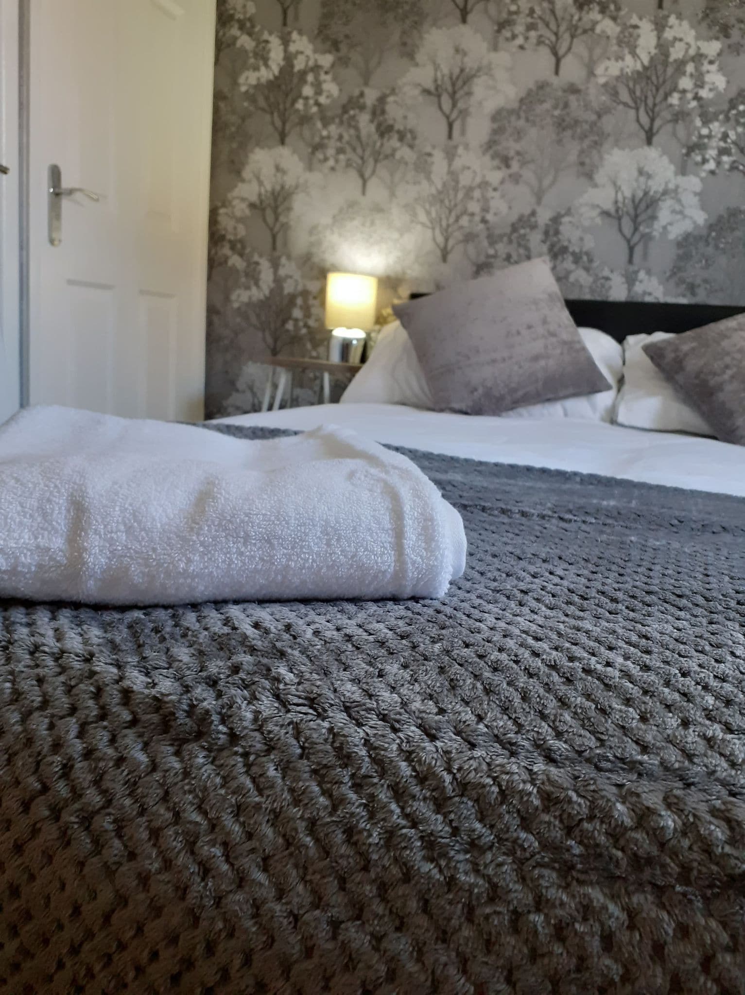 A bed with a textured grey blanket, a white towel, and a patterned wall in the background.
