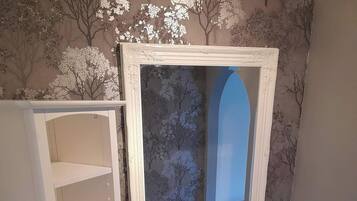 Une salle de bain avec un lavabo sur colonne blanc, un miroir avec un cadre décoratif et un meuble avec plusieurs étagères.