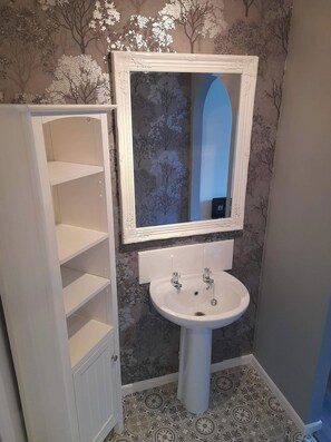 A bathroom with a white pedestal sink, a mirror with a decorative frame, and a cabinet with multiple shelves. - Hardwick Haven, Sedgefield - Near Hardwick Hall (Stockton-on-Tees)