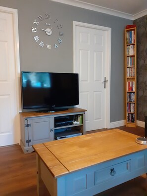 A living room with a television on a wooden stand, a bookshelf, and a wall clock. - Hardwick Haven, Sedgefield - Near Hardwick Hall (Stockton-on-Tees)